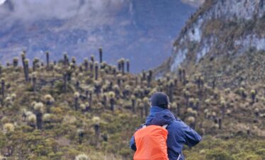 Hiker with orange backpack walking through páramo landscape toward Nevado del Tolima, taking a picture.
