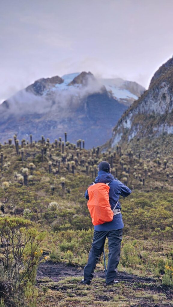 Hiker with orange backpack walking through páramo landscape toward Nevado del Tolima, taking a picture.