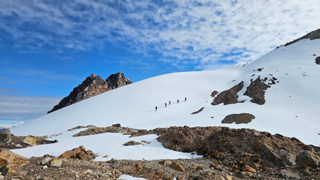 Group of climbers traversing a wide snow slope on Nevado del Tolima in Colombia under a blue sky with scattered clouds.