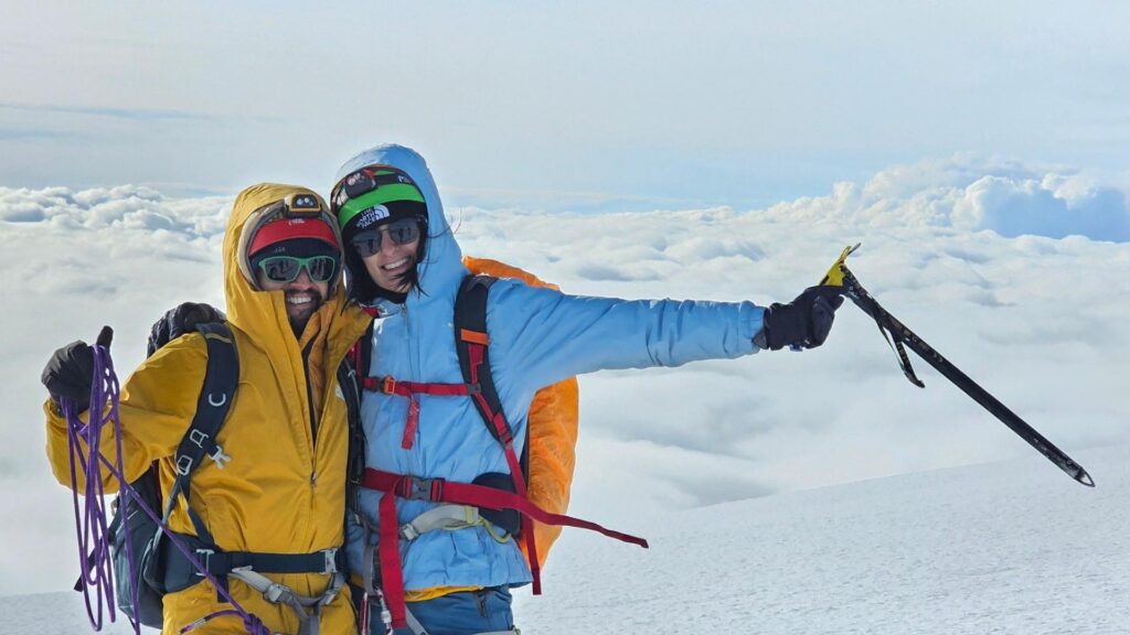 wo climbers in full gear stand on a snowy mountain peak, smiling, with clouds below and a bright sky above. One holds climbing rope, the other an ice axe.