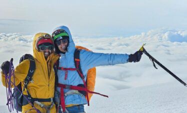 wo climbers in full gear stand on a snowy mountain peak, smiling, with clouds below and a bright sky above. One holds climbing rope, the other an ice axe.