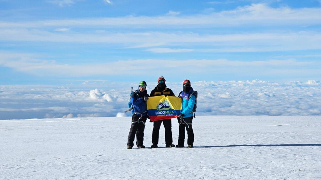 Three climbers on the summit of Nevado del Tolima in Colombia holding a LOCOMBIA flag above the clouds.