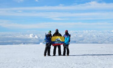 Three climbers on the summit of Nevado del Tolima in Colombia holding a LOCOMBIA flag above the clouds.
