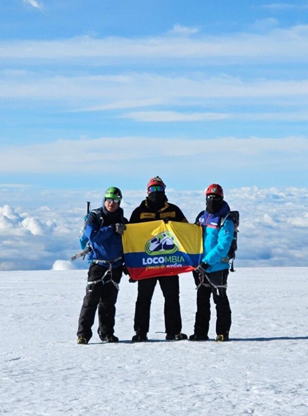 Three climbers on the summit of Nevado del Tolima in Colombia holding a LOCOMBIA flag above the clouds.