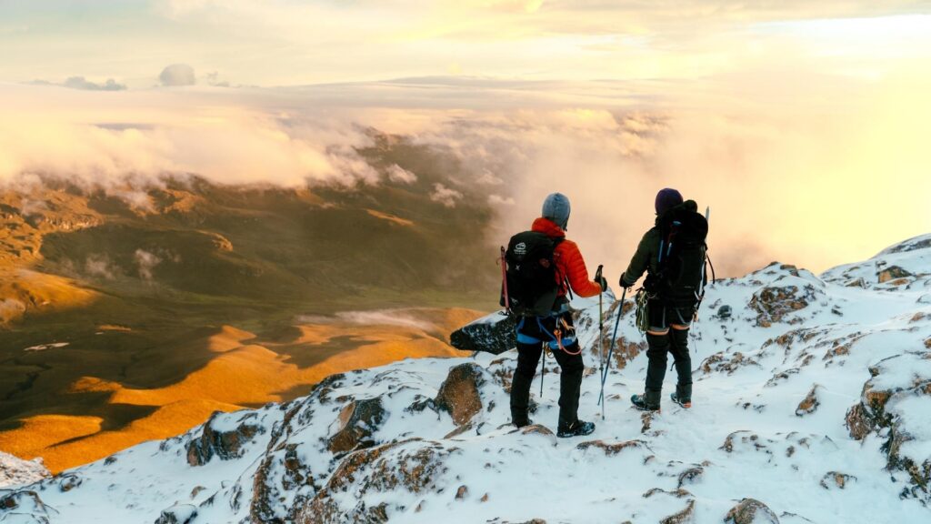 Two climbers standing on the snowy morane of Nevado del Tolima in Colombia, looking over clouds and a sunlit Andean valley at sunrise.