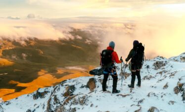 Two climbers standing on the snowy morane of Nevado del Tolima in Colombia, looking over clouds and a sunlit Andean valley at sunrise.