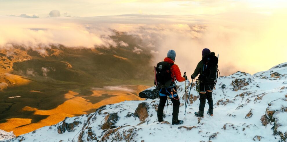 Two climbers standing on the snowy morane of Nevado del Tolima in Colombia, looking over clouds and a sunlit Andean valley at sunrise.