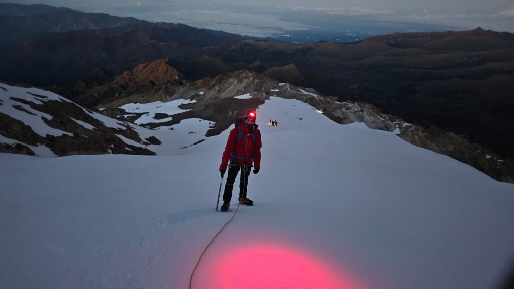 Climber with headlamp ascending Nevado del Tolima before sunrise in Colombia, roped on snowy terrain with mountains in the background.