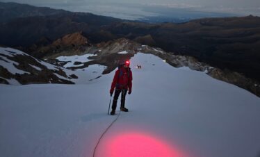 Climber with headlamp ascending Nevado del Tolima before sunrise in Colombia, roped on snowy terrain with mountains in the background.