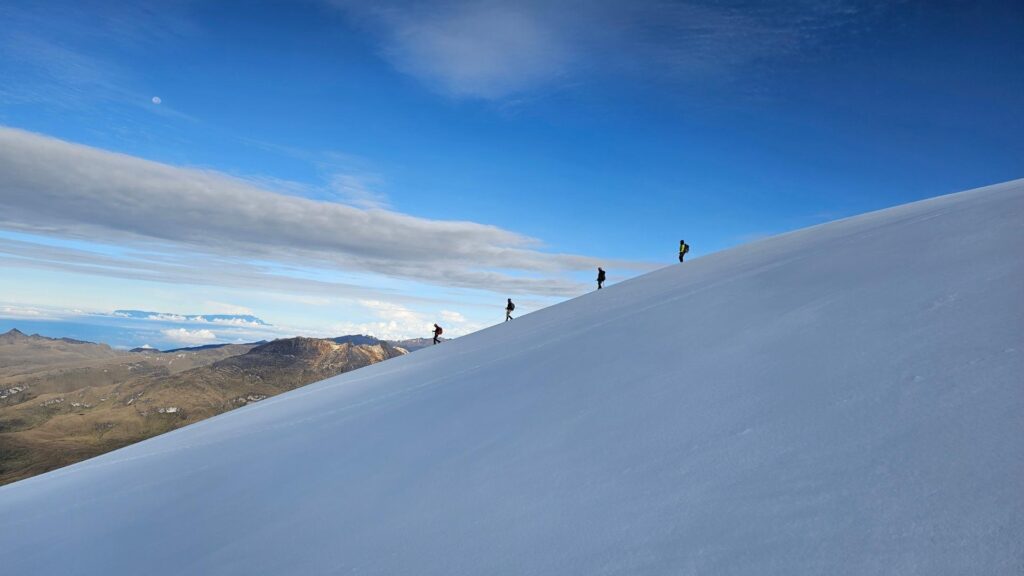 A group of hikers ascend nevado del tolima, snow-covered mountain slope under a bright blue sky with scattered clouds. The moon is visible in the distance.