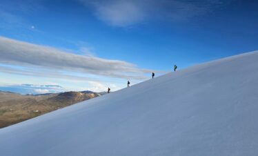 A group of hikers ascend nevado del tolima, snow-covered mountain slope under a bright blue sky with scattered clouds. The moon is visible in the distance.