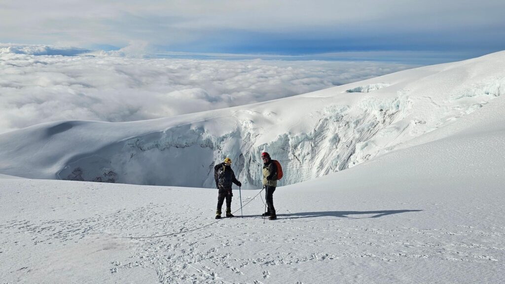 Two hikers stand on the summit of nevado del tolima with clouds below and a glacial ice wall from the volcanic crater behind them.