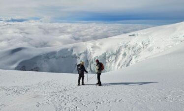 Two hikers stand on the summit of nevado del tolima with clouds below and a glacial ice wall from the volcanic crater behind them.