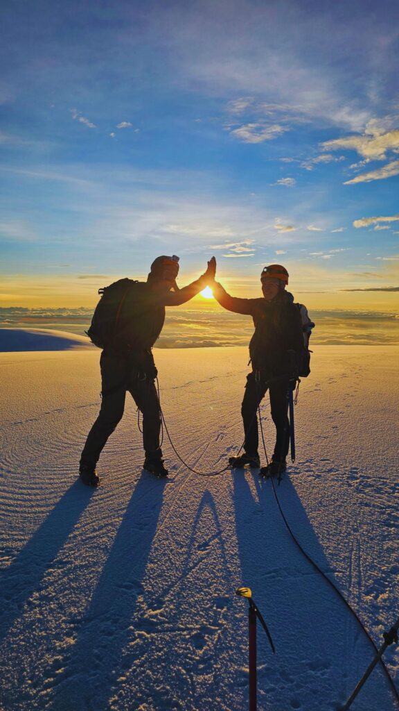 Two climbers high-fiving on the summit of Nevado del Tolima at sunrise above the clouds.