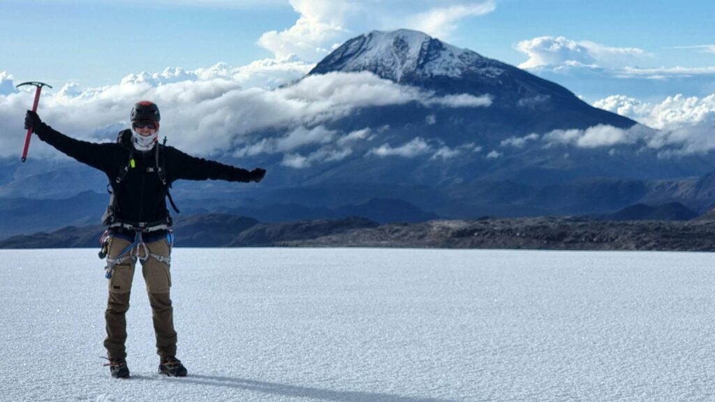 Mountaineer standing on the Nevado Santa Isabel glacier with an ice axe, overlooking Nevado del Tolima volcano rising above the clouds in Los Nevados National Natural Park, Colombia.
