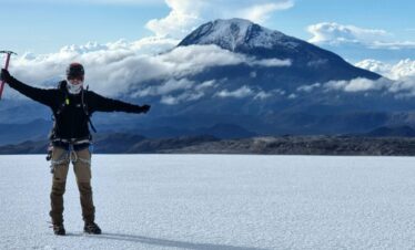 Mountaineer standing on the Nevado Santa Isabel glacier with an ice axe, overlooking Nevado del Tolima volcano rising above the clouds in Los Nevados National Natural Park, Colombia.
