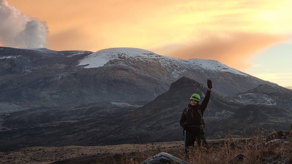 Mountaineer at sunrise on the Nevado Santa Isabel route with Nevado del Ruiz volcano and volcanic plume visible in the background, Colombian Andes.
