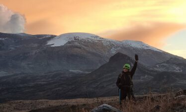 Mountaineer at sunrise on the Nevado Santa Isabel route with Nevado del Ruiz volcano and volcanic plume visible in the background, Colombian Andes.